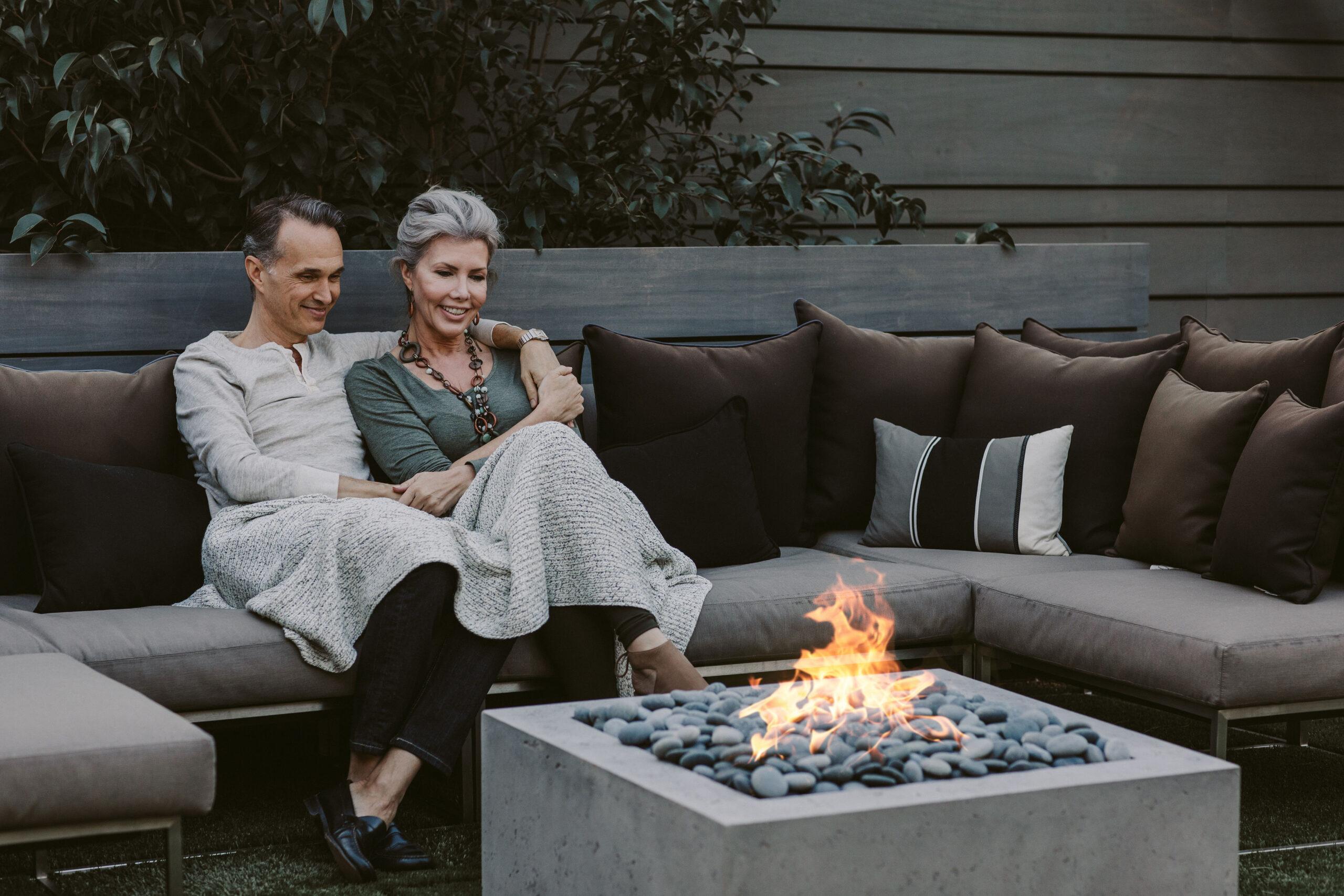 A couple under a blanket sitting on a couch next to a Dekko lightweight concrete fire pit in their backyard.