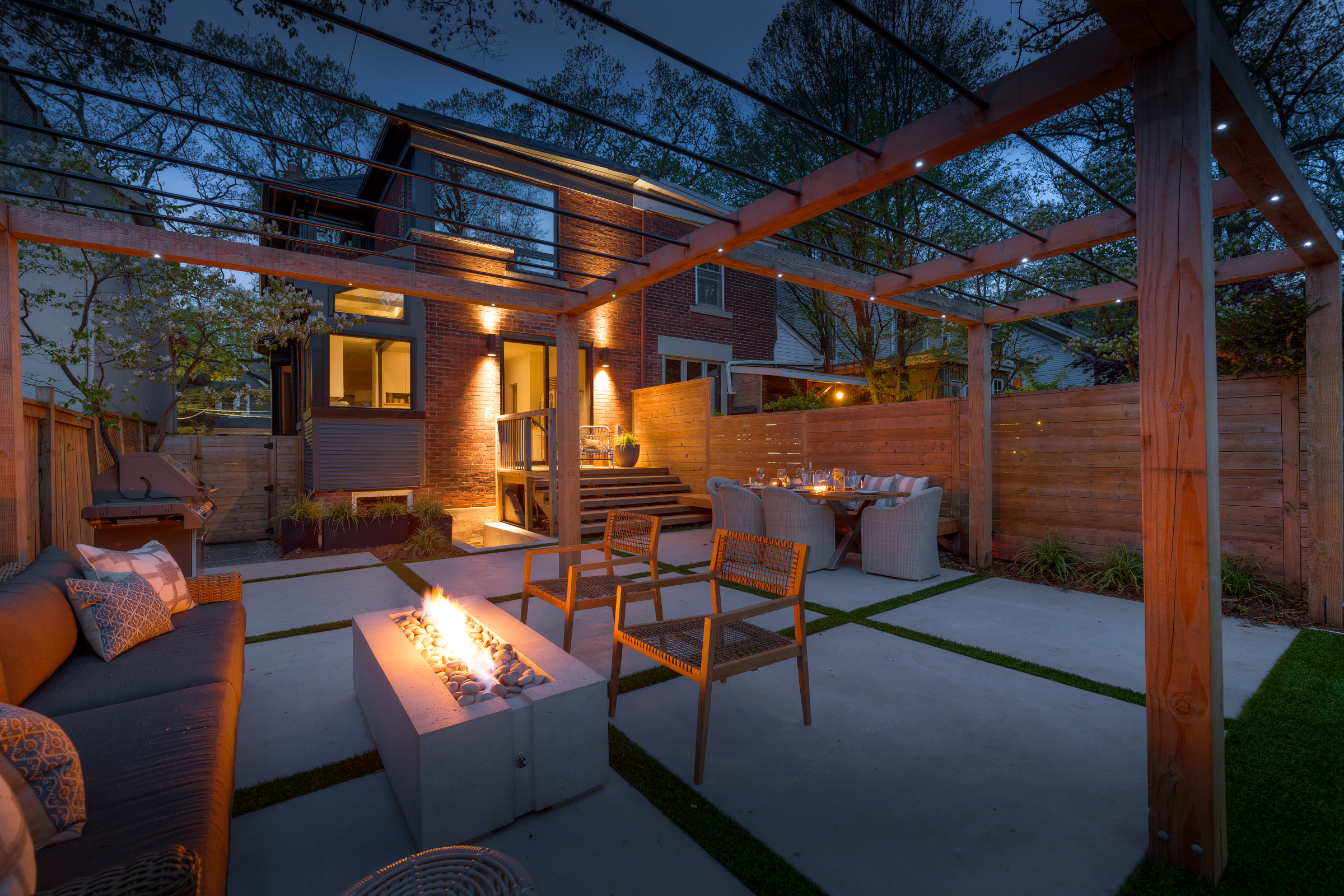 This outdoor space is pictured at dawn, with the rectangular fire pit lit. There is a couch and two chairs next to the fire with a wooden pergola above the seating area.