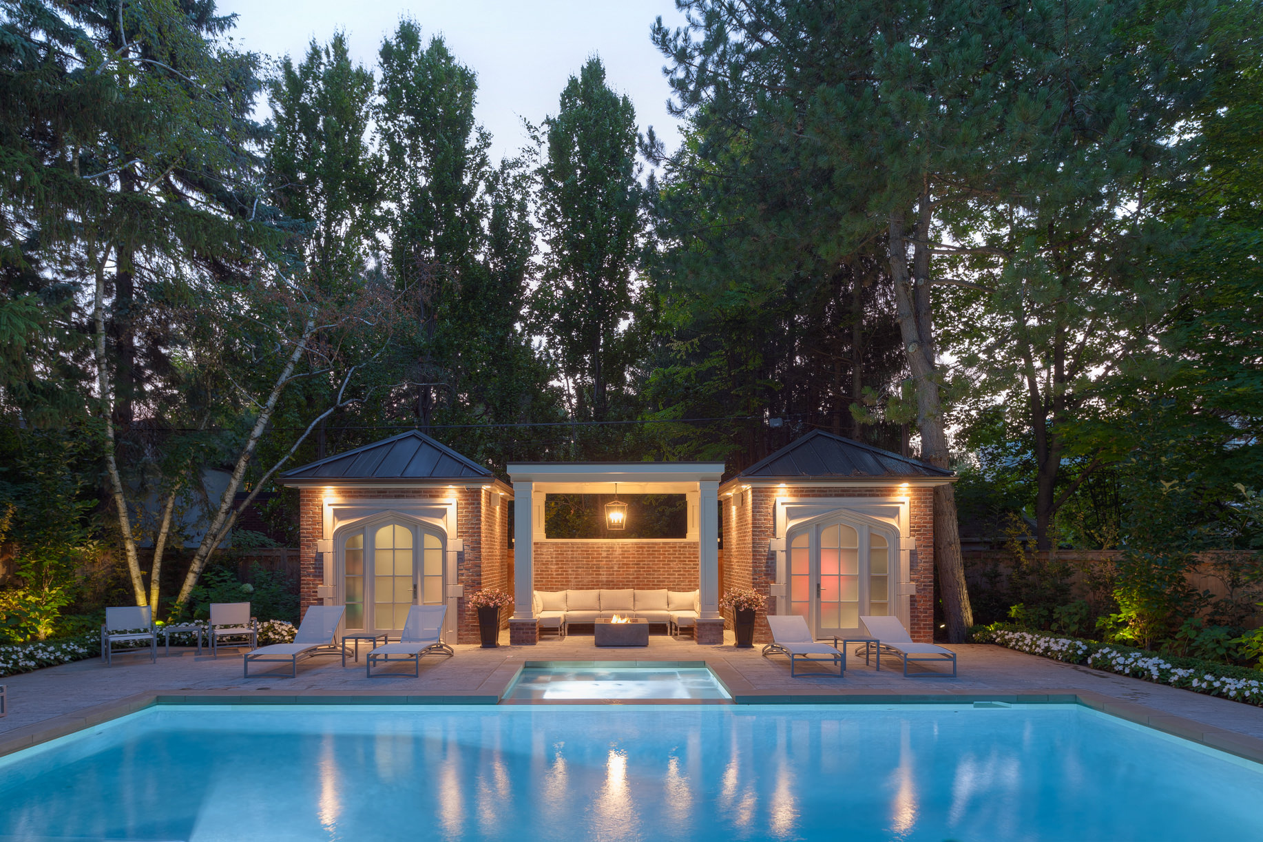 This outdoor space features a large pool and a hot tub. There are two pool houses connected by an outdoor sitting area with a long couch and a firepit.