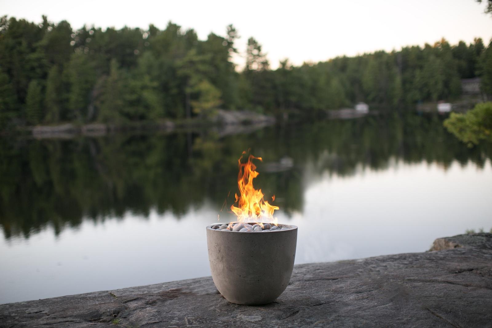 A compact bowl-shaped concrete fire pit from Dekko’s Element collection pictured on a slab of rock in front of a lake at twilight, with a forest in the distance.