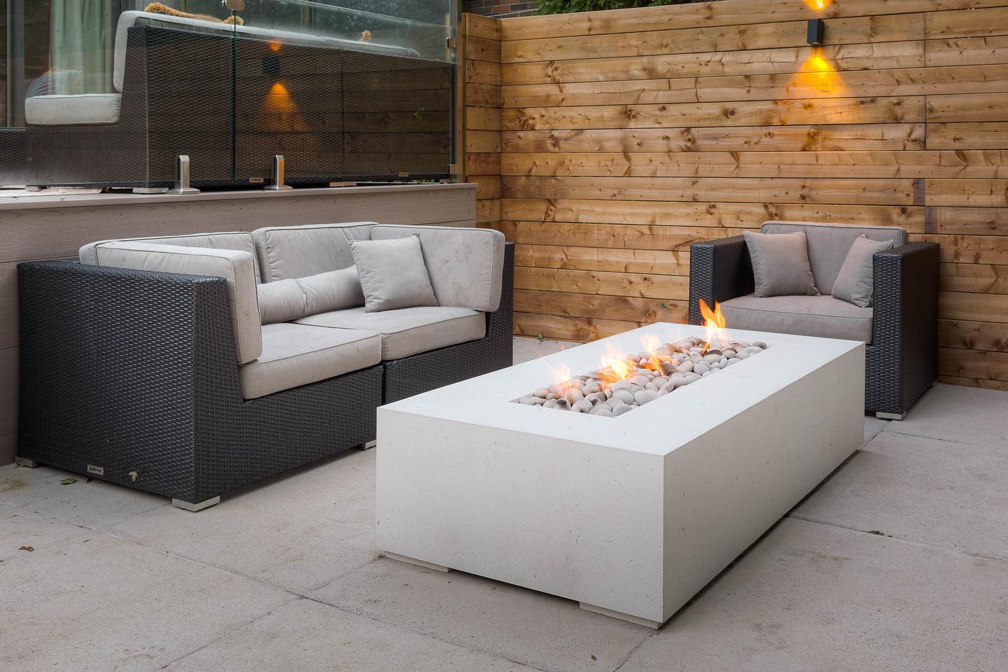 An outdoor concrete patio with a sofa and armchair around a Dekko Sonoma fire pit, lit by a light fixture on a wooden wall.