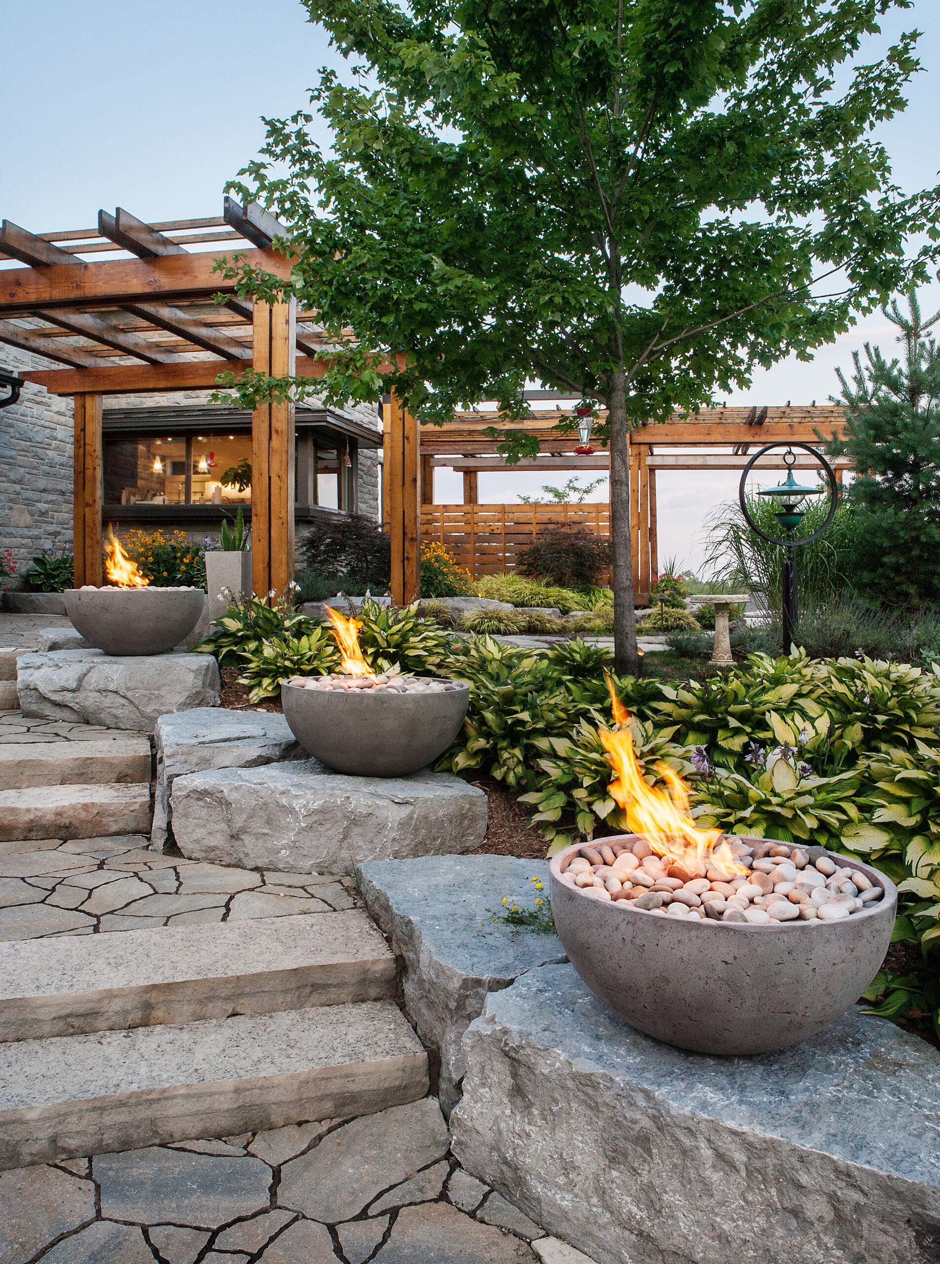 Three Dekko lightweight concrete circular fire pits lining rocks on a walkway to a wooden pergola in a backyard.
