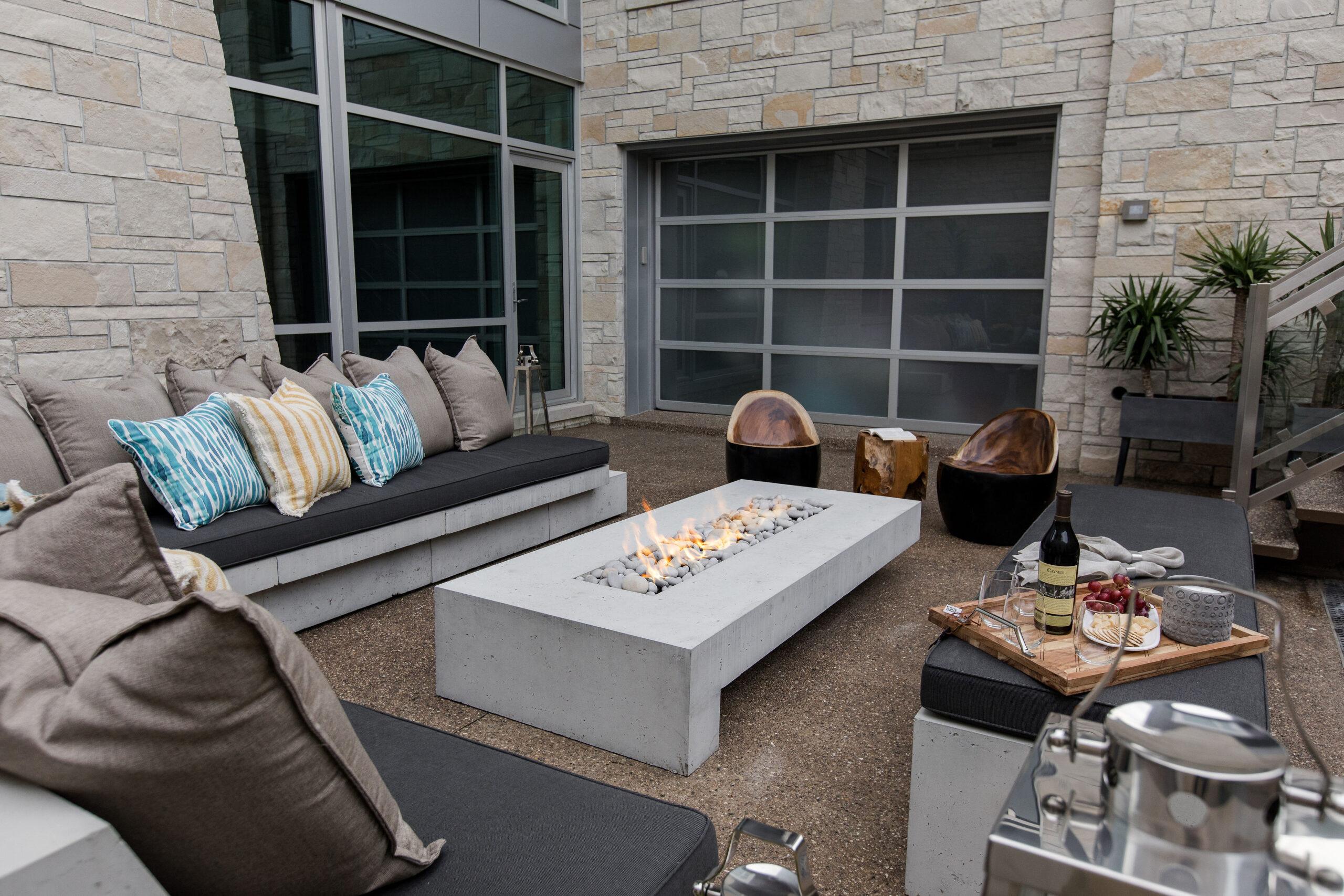 A rectangular Dekko lightweight concrete fire pit in a backyard with couches, wooden chairs, and a cheese board surrounding it.